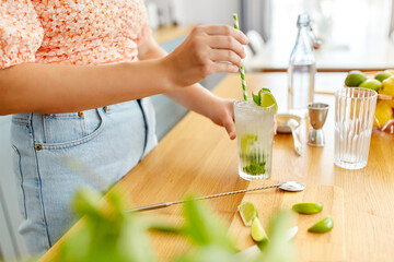 drinks and people concept - close up of woman with paper straw making lime mojito cocktail at home kitchen