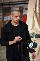 caucasian young handsome man gafer with an earring in his ear and a white hairpiece adjusts the power of the lighting lantern on the movie set