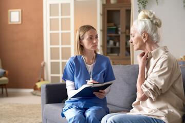 Senior woman complaining about her health to nurse while she making notes in card, they sitting on...
