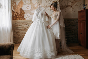 Fototapeta premium Blonde Bride in a robe, posing for the camera looking at her wedding dress. Elegant hairstyle. Diadem. Beautiful makeup. Morning of the bride