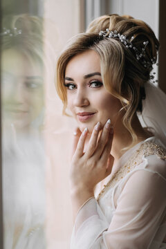 Blonde bride with elegant hairdo and tiara, wearing a robe, posing, looking at the camera, touching her face. Beautiful makeup. Elegant hairstyle. French manicure