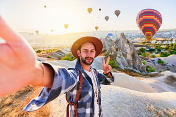 Happy bearded guy traveling blogger with victory sign takes selfie photo in Nevsehir, Goreme. Beautiful destination with flying hot air balloons in Anatolia, Kapadokya