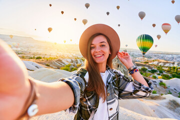 Happy smiling joyful satisfied girl traveling blogger takes selfie photo in Nevsehir, Goreme. Beautiful destination with colorful flying hot air balloons in Anatolia, Kapadokya
