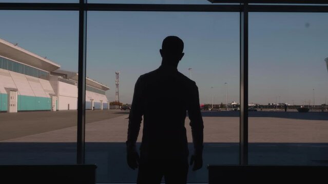 A man walking down a corridor with large windows. HDR. A man in a shirt and jeans walks through the office where a beautiful evening sunset is visible.
