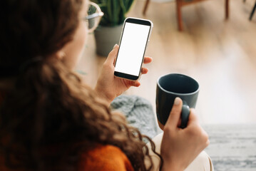 Over-shoulder view of woman holding coffee and smartphone with white mock-up cellular screen in hands using mobile apps on a cell phone. Cellphone display mock-up for advertising concept.