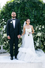 beautiful newlyweds on the background of an arch with greenery and flowers holding hands