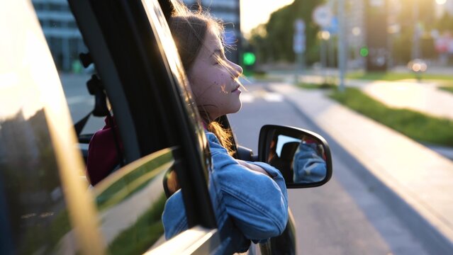 Happy Caucasian girl enjoying looking out the window in car traveling in city on road trip vacation. Little pretty child sits in auto pulling her face out of car window on sunny day. Close up concept
