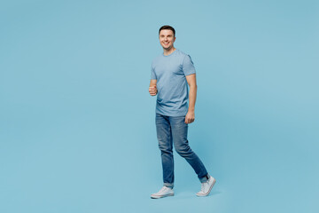 Naklejka premium Full body side view young satisfied smiling happy man wears casual t-shirt walking strolling going look camera isolated on plain pastel light blue cyan background studio portrait. Lifestyle concept.