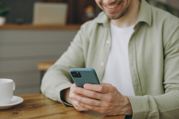 Cropped close up young fun caucasian man wear casual clothes sits alone at table in coffee shop cafe restaurant indoors work hold use mobile cell phone look aside rest relax during free time inside.