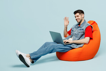 Full body fun young IT man wear denim vest red t-shirt casual clothes sit in bag chair hold use work on laptop pc computer waving hand isolated on plain light blue background studio Lifestyle concept