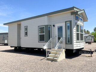 A manufactured tiny home sits on a gravel lot with wheels and temporary stairs, illustrating...