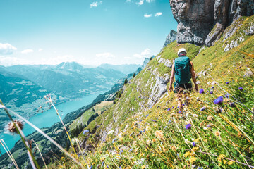 Back view of mountaineer hiking on flowery trail next to steep rock wall with panoramic view on Walensee and the swiss alps in the background. Schnürliweg, Walensee, St. Gallen, Switzerland, Europe. - obrazy, fototapety, plakaty