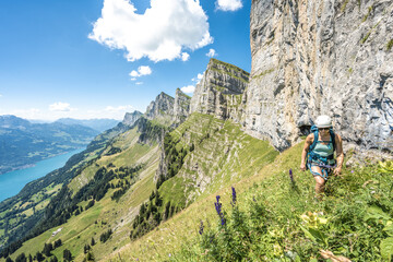 Athletic woman hiking on flowery meadow below steep rock wall with scenic view on lake Walensee and...