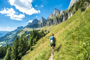 Fototapeta na wymiar Sporty woman walks on scenic hiking trail between meadow and trees and the Churfürsten mountain range in the background. Schnürliweg, Walensee, St. Gallen, Switzerland, Europe.