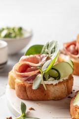 Bruschetta with ham or prosciutto, cream cheese, cucumber, spinach and microgreens. Tasty sandwich or toasted bread on white tray close up