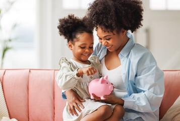 financial education. african american family, mother and child daughter with pig piggy bank counting savings at home  . - obrazy, fototapety, plakaty