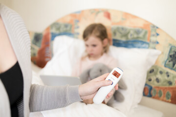 mother measuring temperature of her daughter. Kid sitting in bed with tablet and furry toy and no face woman holding infrared thermometer which indicates 38,2 C