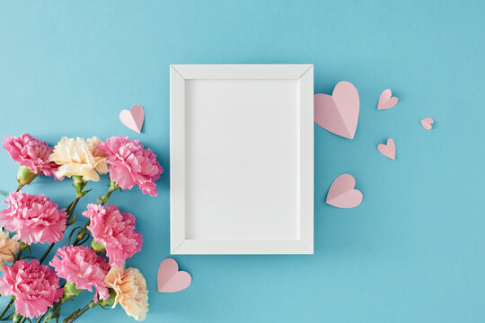 Charming Mother's Day flower gift concept. Top view photo of pretty carnations flowers and paper hearts on light blue background. Flat lay with empty frame for greeting text or advert