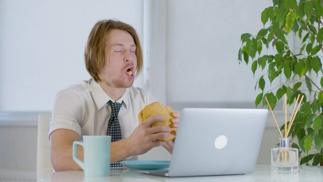 red-haired young male worker in the office.eating a burger in tasty laptop