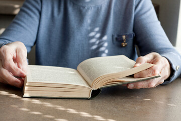 Closeup of elderly woman hand sitting at table reading an fiction book. Old lady spending time reading paper book at home. Reading helps to train brain activity.