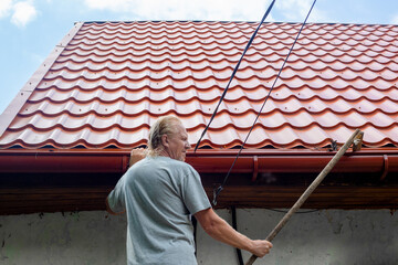 A mature man cleans the gutter of a drainpipe on the roof of his house from autumn leaves and debris.