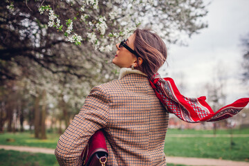 Back view of stylish woman wearing red hair scarf in spring park. Retro female fashion. Headscarf for bun hairstyle - obrazy, fototapety, plakaty