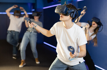 Fototapeta na wymiar Cheerful emotional teenager in VR headset fully immersed in game, actively engaged in virtual reality world, manipulating objects or shooting targets with joysticks in hands..
