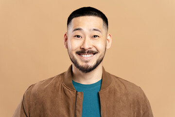 Obraz premium Portrait of confident smiling asian man with stylish beard, hairstyle wearing brown jacket isolated on beige background. Happy attractive Korean fashion model looking at camera, studio shooting 