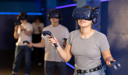 Cheerful emotional young woman in VR headset fully immersed in game, actively engaged in virtual reality world, manipulating objects or shooting targets with joysticks in hands..