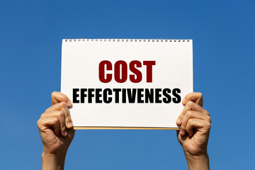 Cost effectiveness text on notebook paper held by 2 hands with isolated blue sky background. This message can be used as business concept about cost effectiveness. - obrazy, fototapety, plakaty