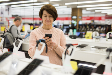 Mature caucasian woman choosing smartphone in electronic shop.