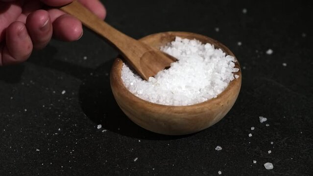A girl dials salt into a wooden spoon from a bowl on a black background. Coarse sea salt.