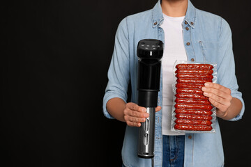 Woman holding sous vide cooker and sausages in vacuum pack on black background, closeup. Space for...