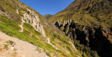 A narrow path along the steep slope of a high mountain with unusual sandstone rock formations at the top.