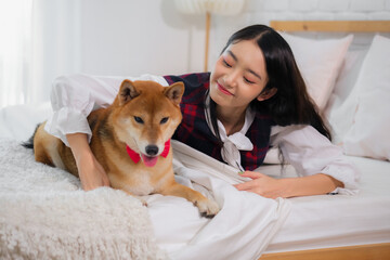 Beautiful asian woman lying on a white bed in the bedroom while embracing a dog with joy expression.