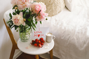 Breakfast for Mothers Day. Heart shaped white plate with fresh strawberries, cup of coffee, gift and Peonys bouquet with gift in bed. Still life composition. Happy Mother's Day.