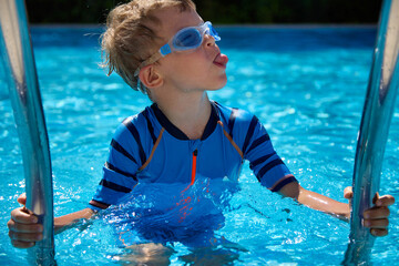 Funny boy in a blue bathing suit stands on a step in the pool, holding on to a metal railing, shows...