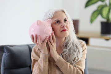 Senior woman puts ear to piggy bank sitting in armchair