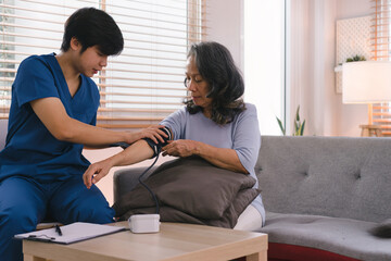 Asian caregiver and doctor examine older patient using blood pressure gauge. A young Asian male therapist and nurse are taking care of a senior elderly woman sitting on a sofa. - Powered by Adobe