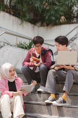A multiethnic group of university students is studying together while sitting on steps near the campus and communicating with each other.