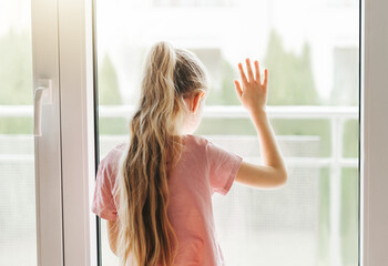 Sad little girl looking through  window at home.