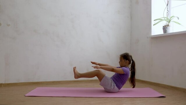 School age girl sitting keep raised bent legs on exercise mat in empty room, then look at camera and smile, side view, free copy space. Home workout, gymnastics, kids sport, balance, press, activity.