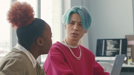 Young Asian blue haired male programmer chatting with African American female colleague while program coding on laptops at desk in office - Powered by Adobe