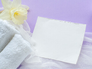 Daffodils flower, towels and paper card with copy space on lilac background. Women and Mothers Day gift. Concept of wellness, SPA and relaxation. Congratulations note and towels, white violet flat lay
