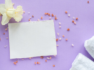 Daffodils flower, white towels, sea salt and paper card with copy space on lilac background. Women and Mothers Day gift. Concept of wellness, SPA and relaxation. Note and towels flat lay