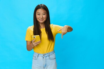 Happy Asian woman holding a phone with a yellow case on a blue background in a yellow T-shirt with a smile and teeth pointing her finger at