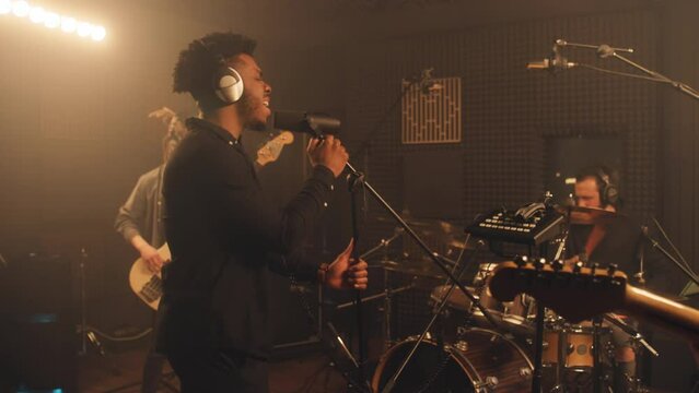 Cool multi ethnic rock band in process of creating new music album in sound recording studio. African American singer keeps hands on microphone stand and sings. Soundproof room. Music production.