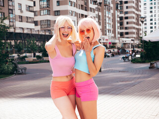 Two young beautiful smiling hipster female in trendy summer clothes. Sexy carefree women posing in the street in wigs. Positive funny models having fun at sunset. In sunglasses. At sunny day