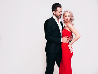 Beautiful fashion woman and her handsome boyfriend in black elegant suit. Sexy blond model in red evening dress. Fashionable couple posing in studio. Brutal man and his female. Isolated on white