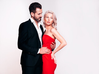 Beautiful fashion woman and her handsome boyfriend in black elegant suit. Sexy blond model in red evening dress. Fashionable couple posing in studio. Brutal man and his female. Isolated on white
